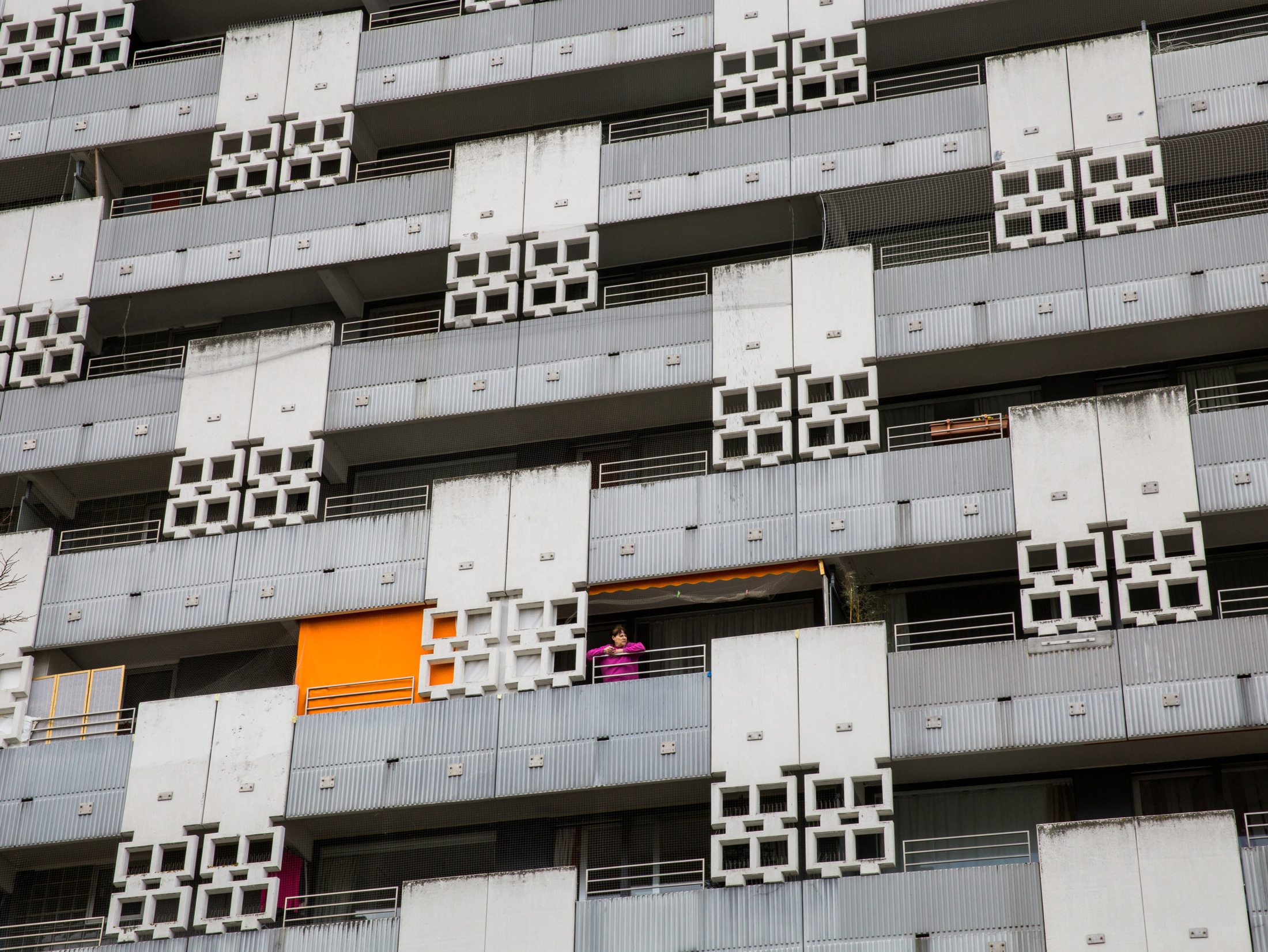 graue Häuserfassade mit Balkonen und einem orangenen Vorhang. Eine Frau steht auf einem der Balkone und sieht nach rechts