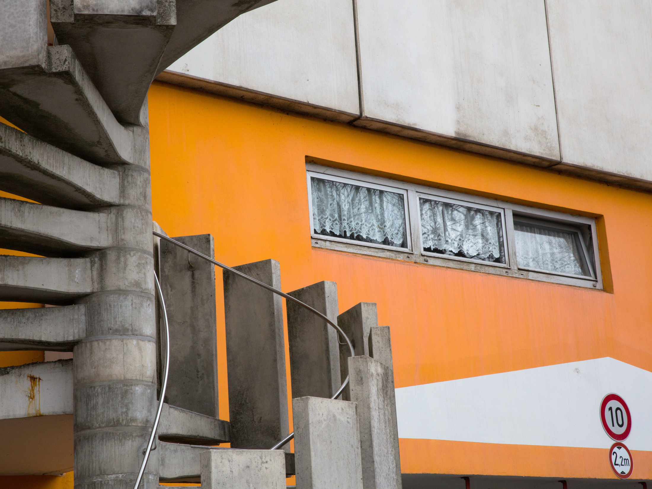 eine orangene Fensterwand hinter einer engen Beton-Wendeltreppe