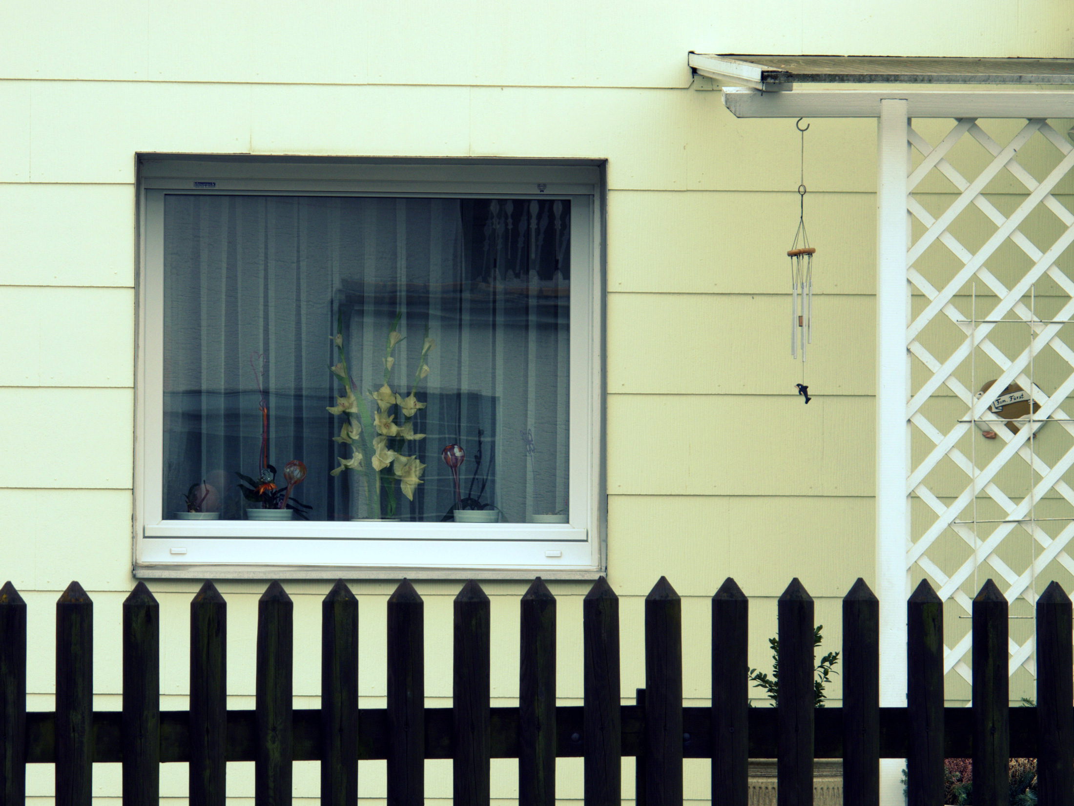 ein Fenster mit Blumen vor den Gardinen und ein Windspiel am Eingangsbereich